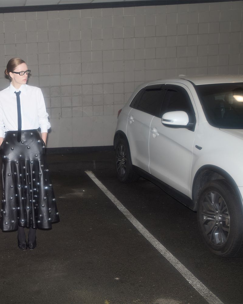 Adult female with glasses and tie stands in parking garage next to white SUV. She wears a studded black skirt and white shirt.