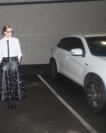 Adult female with glasses and tie stands in parking garage next to white SUV. She wears a studded black skirt and white shirt.
