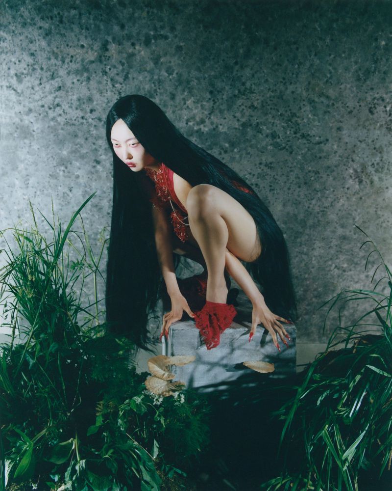 Young Asian woman with long black hair poses crouching on a rock wearing red outfit amidst lush green foliage against a mottled wall.
