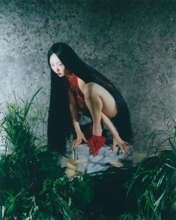 Young Asian woman with long black hair poses crouching on a rock wearing red outfit amidst lush green foliage against a mottled wall.