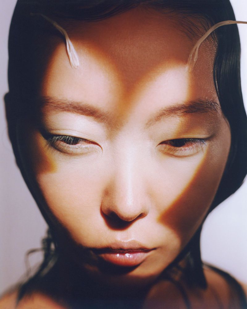 Close-up beauty shot featuring an Asian female model with an updo hairstyle, dramatic lighting, and subtle feather accessories.