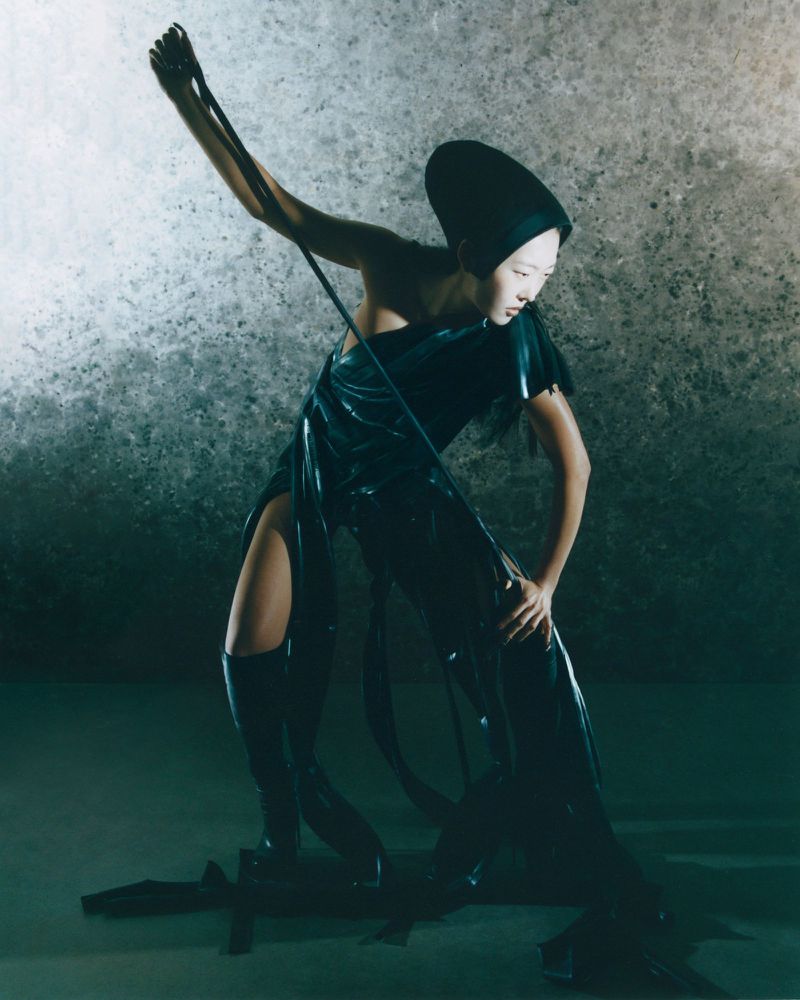 Model Hey Yeon in a sculpted black draped outfit and headpiece posing dynamically against a textured grey studio backdrop.