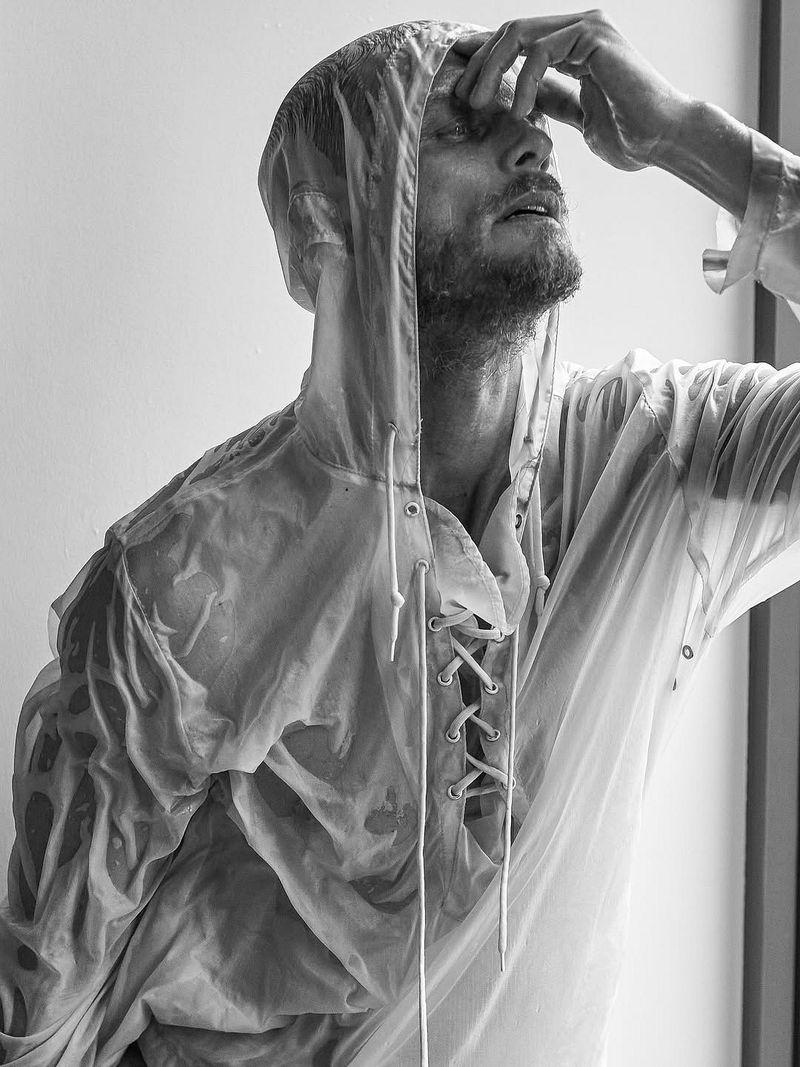 Black and white portrait of a man wearing a transparent hooded jacket, his hand on his forehead, gazing up in a brightly lit indoor setting.