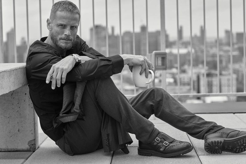 Monochrome portrait of a mature man in a dark shirt, seated outdoors holding a mug, with a cityscape in the background.
