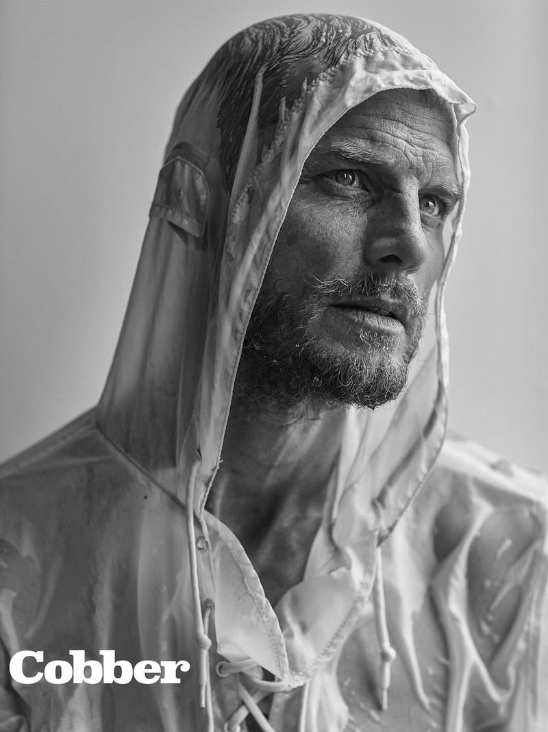 Black and white portrait of a bearded adult male wearing a translucent hooded raincoat, looking off-camera in an indoor environment.