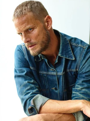 Man with short blonde hair and beard wearing a blue denim jacket against a bright white background