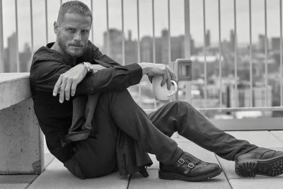 Arnaud Lemaire seated on a rooftop holding a white mug, wearing a dark shirt, trousers, and double-monk strap shoes against a city skyline.