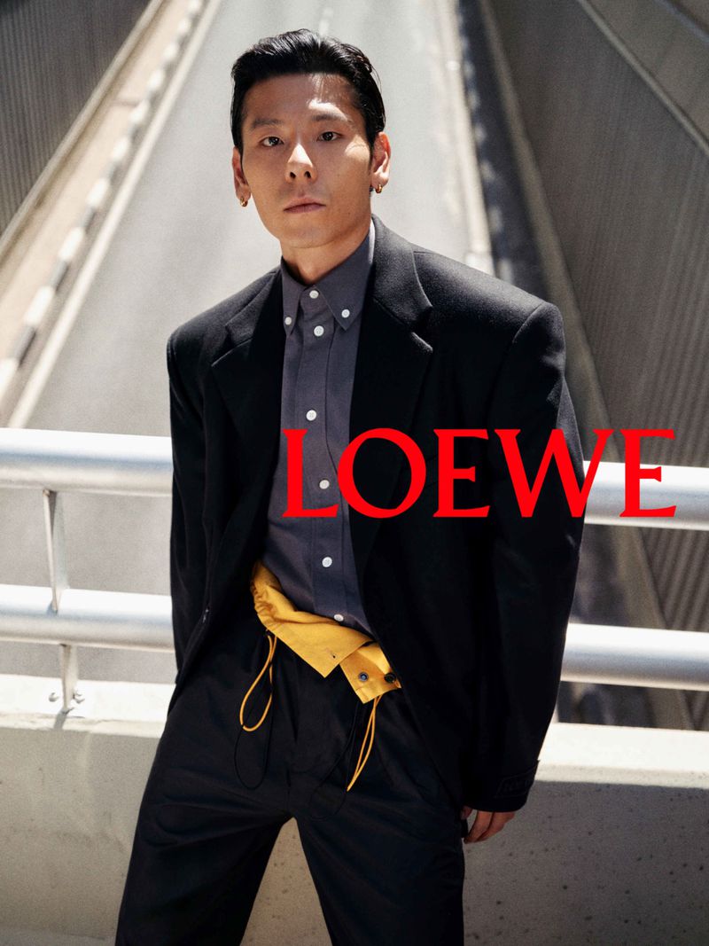 Man wearing a black blazer and grey shirt posing against a bridge railing for Loewe pre collection campaign