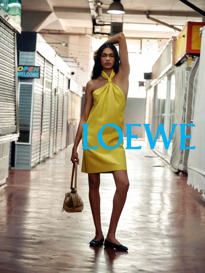 Model in a yellow halter dress and flat shoes holding a tan handbag inside a covered market arcade