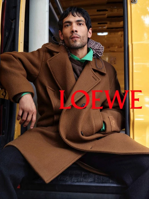 Man wearing a brown coat and green collar seated in front of a yellow vehicle with red Loewe text overlay