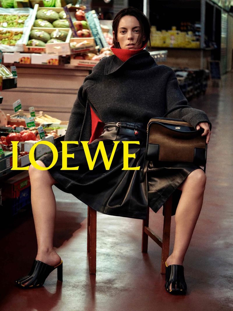 Woman seated on a chair in a market holding a handbag, wearing a dark sweater and leather skirt.