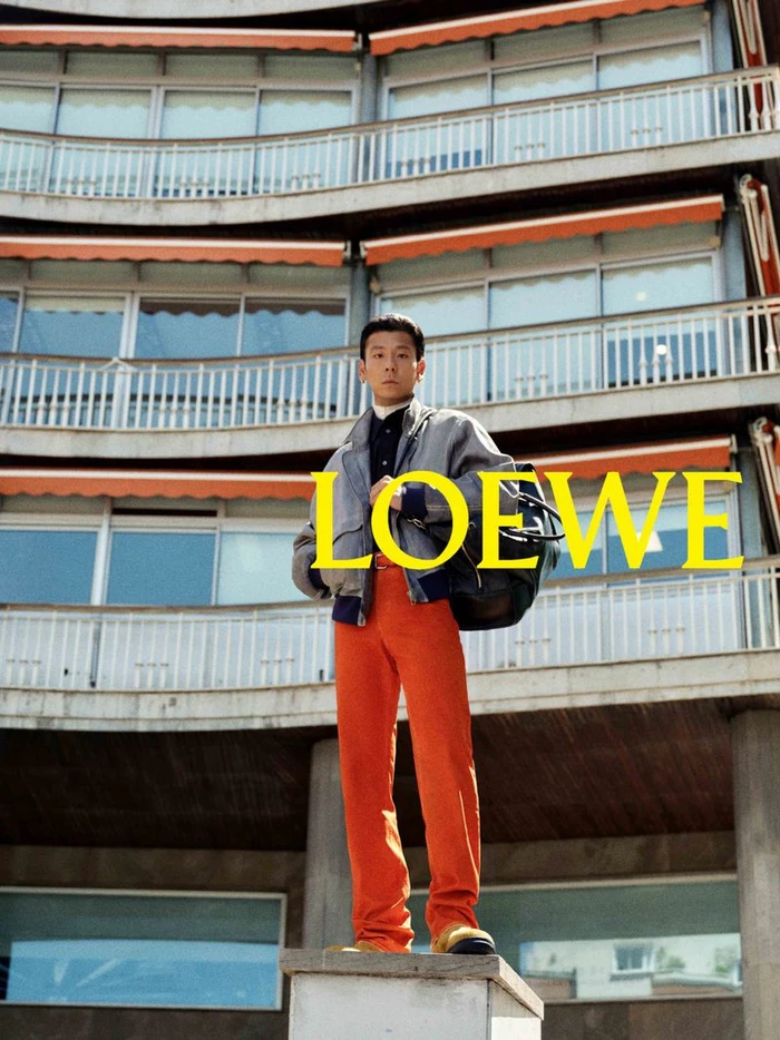 Man standing on a concrete structure in front of a curved multi-story building wearing orange trousers and a gray jacket