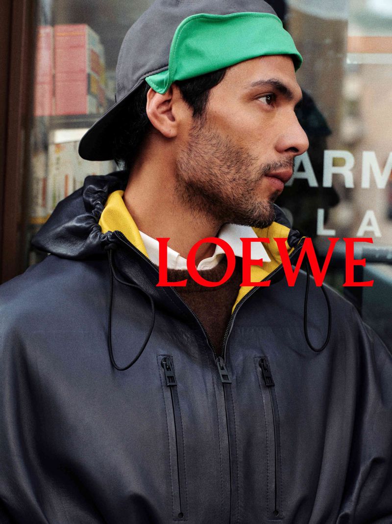 Man wearing a dark leather jacket, layered yellow hoodie, and a grey cap with a green brim for Loewe S/S 27 Pre Collection Campaign.