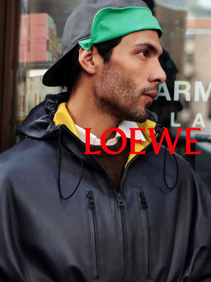 Man wearing a dark leather jacket, layered yellow hoodie, and a grey cap with a green brim for Loewe S/S 27 Pre Collection Campaign.