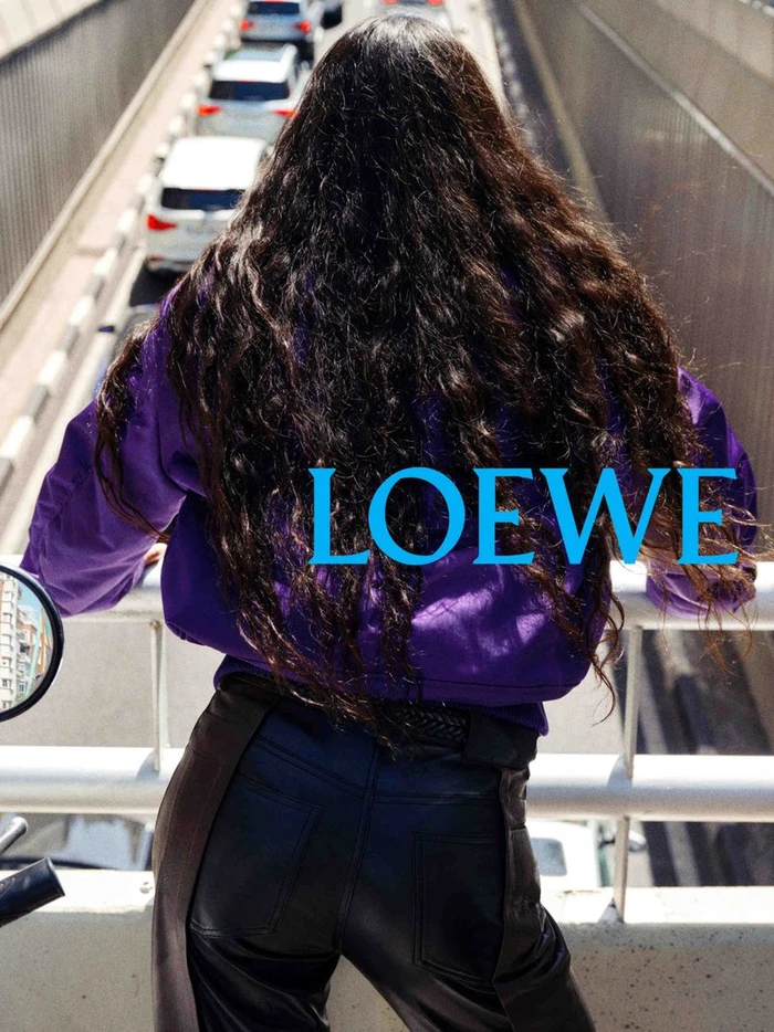 Rear view of a person with long dark hair wearing a purple jacket looking down from a bridge over traffic