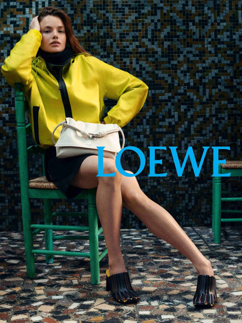 Model in bright yellow jacket and black mules sitting on a green chair holding a white handbag for Loewe