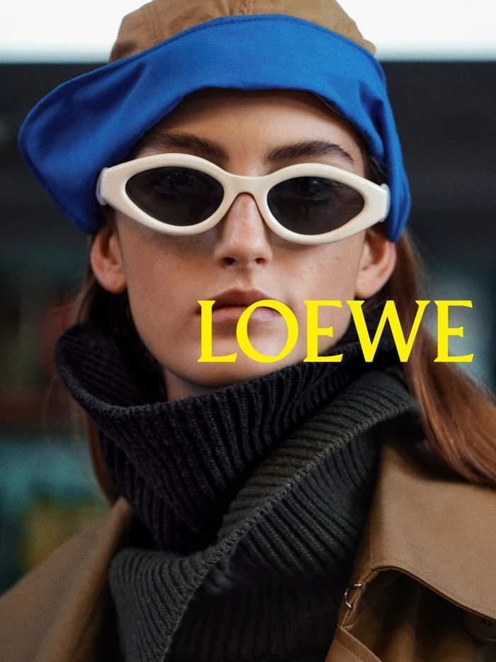 Model wearing white oval sunglasses, a blue and brown cap, and a dark ribbed turtleneck in a Loewe pre-collection campaign image.