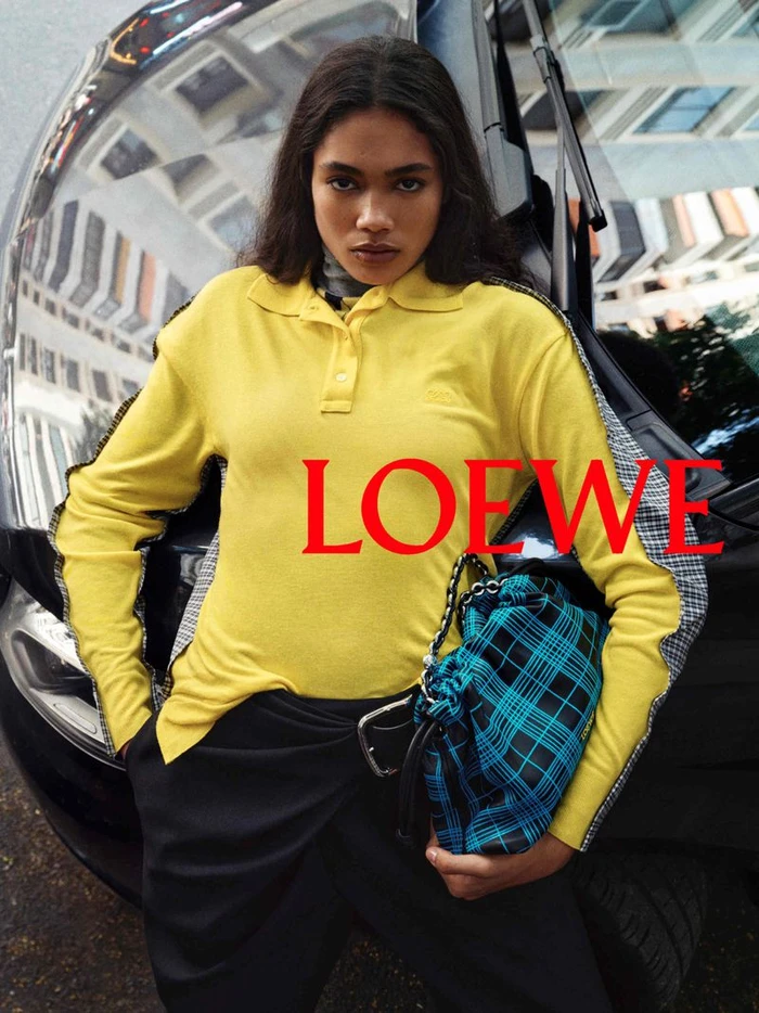 Model in yellow polo shirt leaning against a black car holding a blue plaid handbag for Loewe