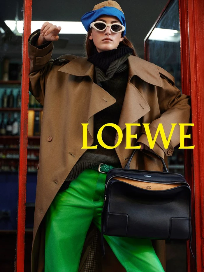 Model in brown trench coat, green pants and blue cap holding a black handbag in a doorway