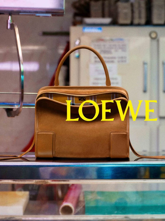 Brown leather handbag centered on a reflective glass shelf with bright yellow text overlaid reading Loewe