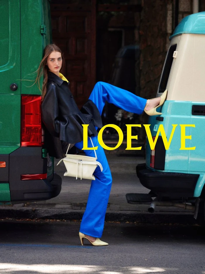 Model leaning against a green van and blue van wearing a black jacket and blue trousers holding a white handbag.