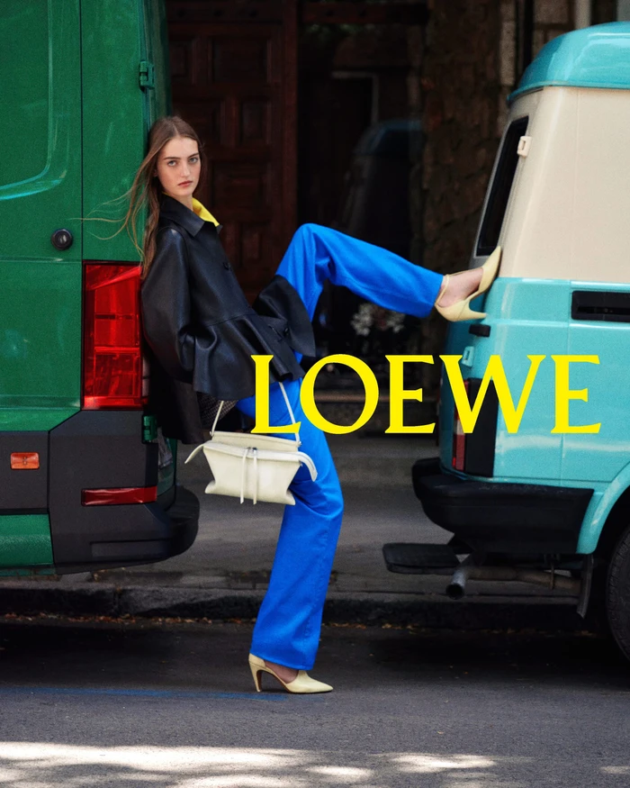 Model in a black jacket and blue trousers leaning between a green van and a turquoise van on a street