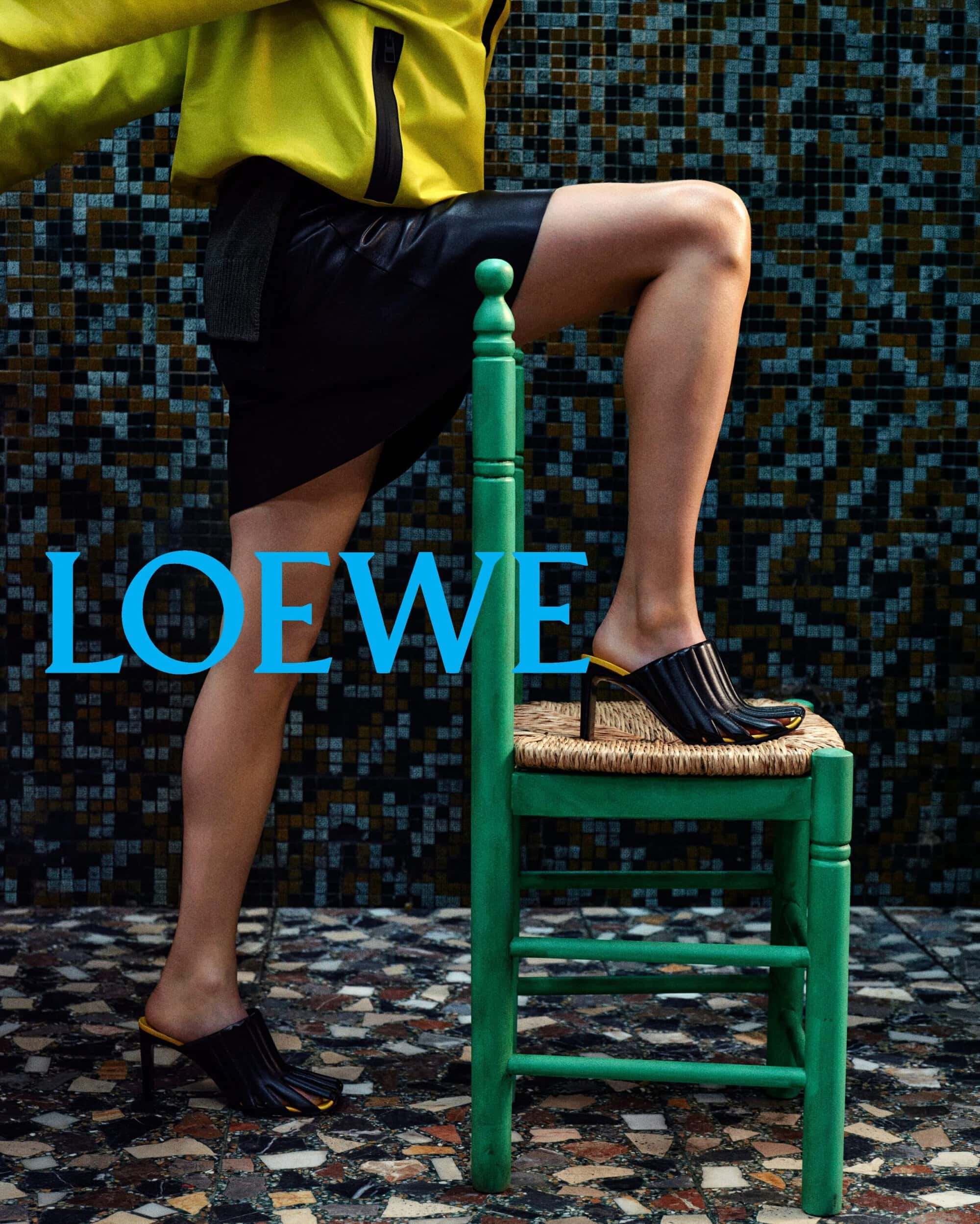 Model wearing black heeled mules resting a leg on a green chair against a tiled wall in a Loewe campaign image
