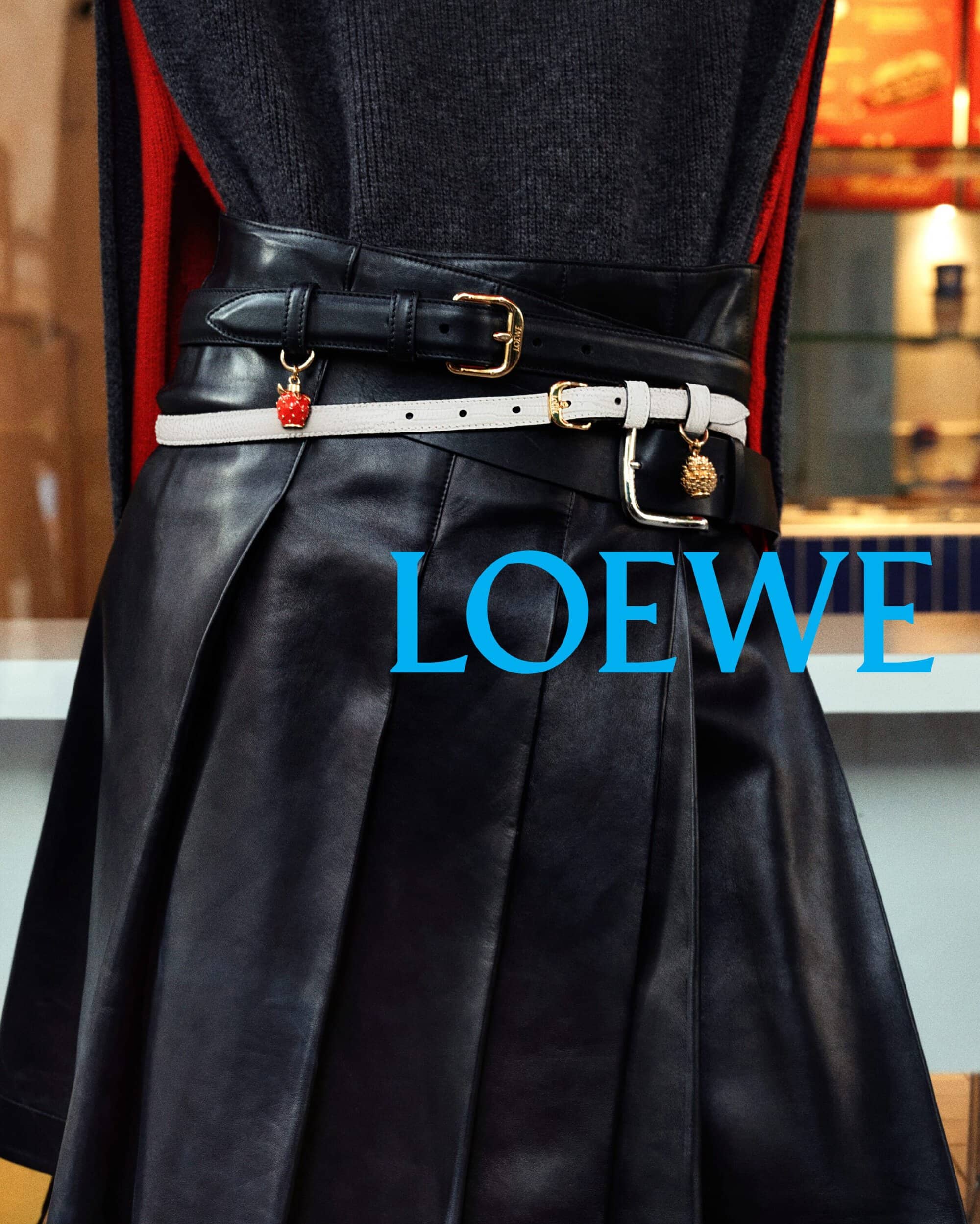 Close-up of a person wearing a black pleated leather skirt cinched with two thin belts, featuring hanging fruit charms and the Loewe logo.