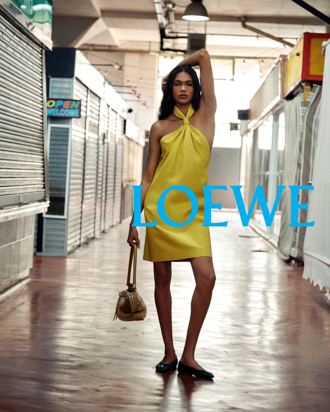 Model in a halterneck yellow dress holding a handbag in a market arcade with Loewe overlay text