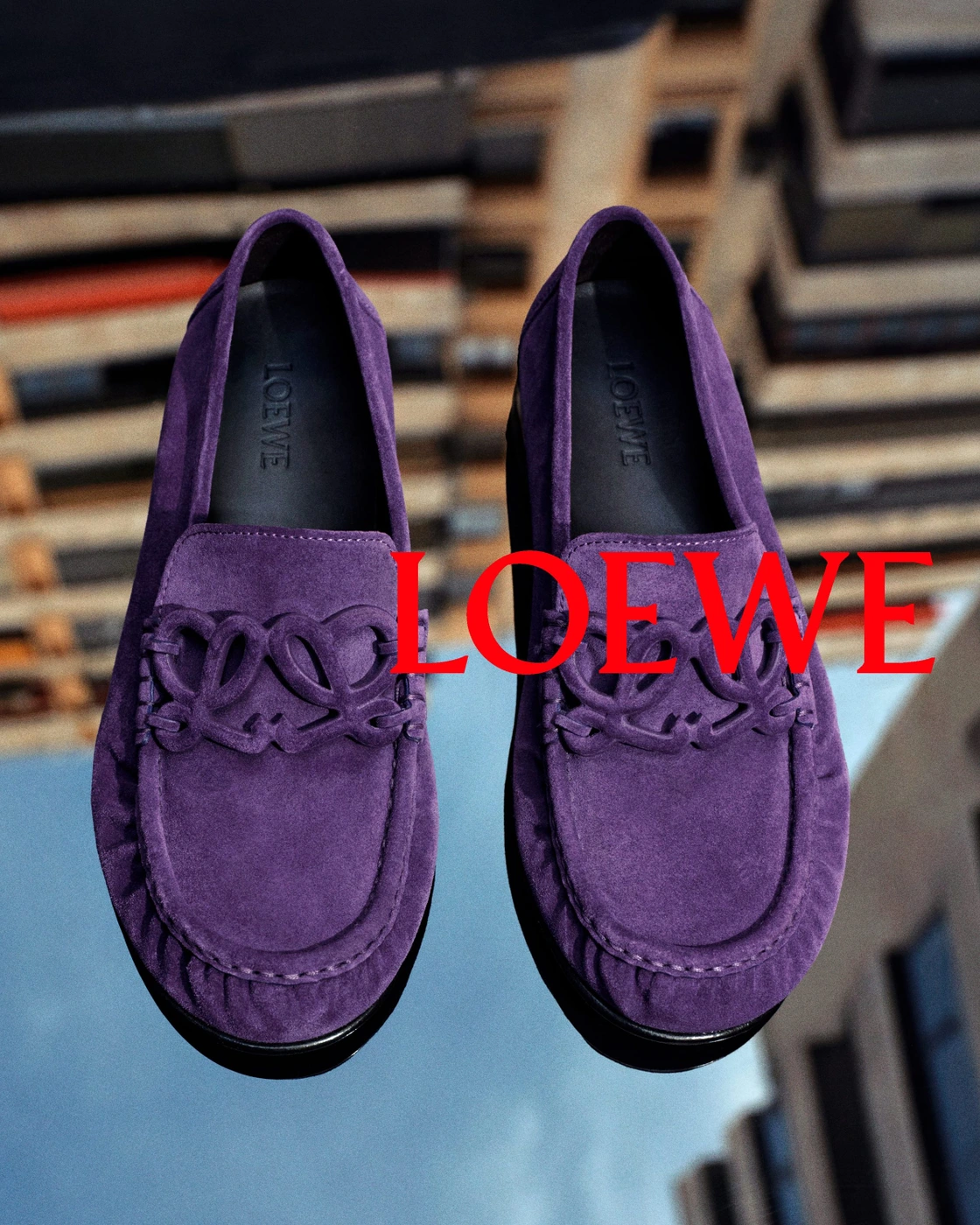 Pair of purple suede loafers with cutout anagram strap details positioned vertically against a blurred urban background.