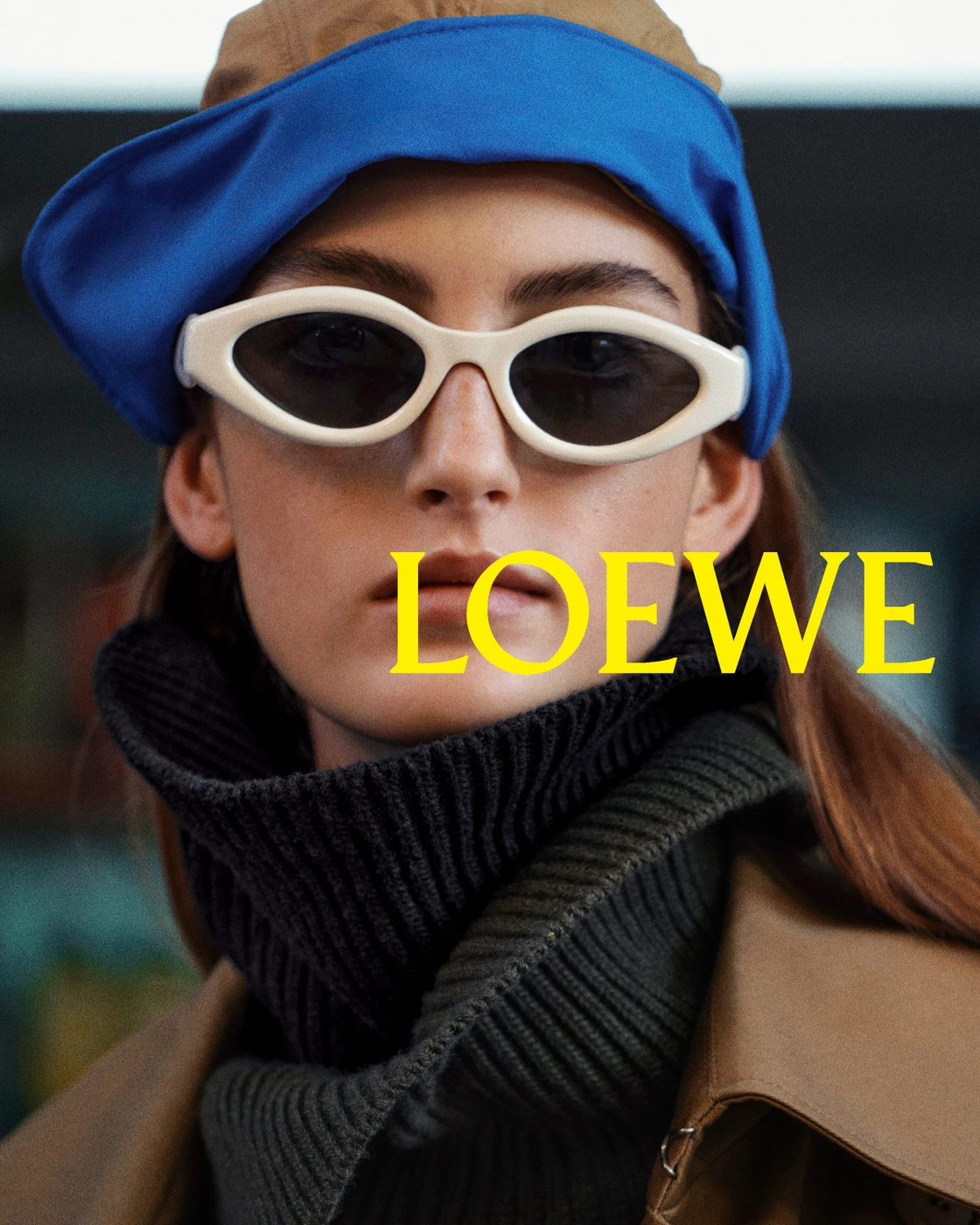 Close-up portrait of a model wearing white sunglasses, a blue cap, dark turtleneck, and brown jacket for Loewe campaign.
