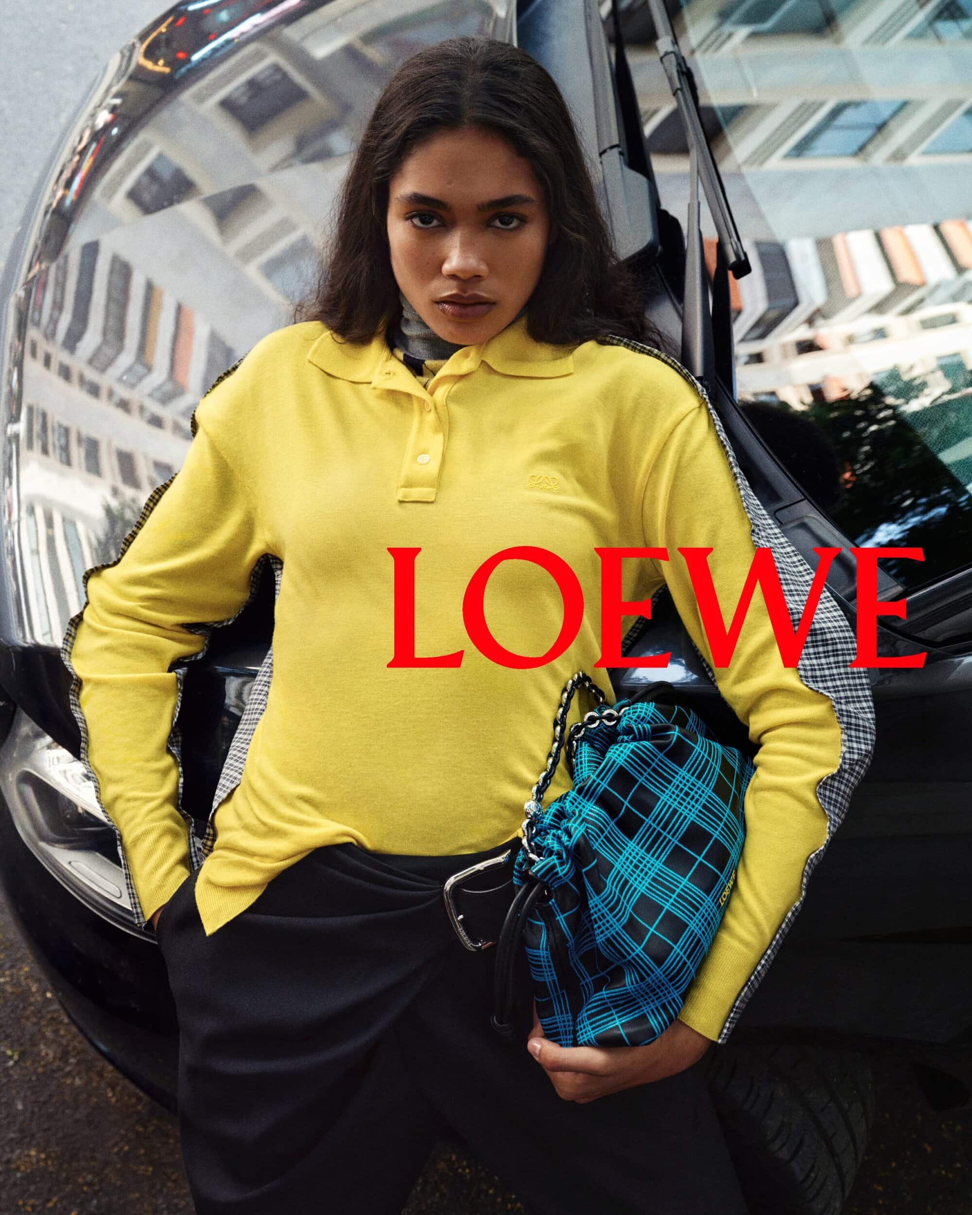 Model in yellow polo shirt leaning against a black car holding a blue plaid handbag