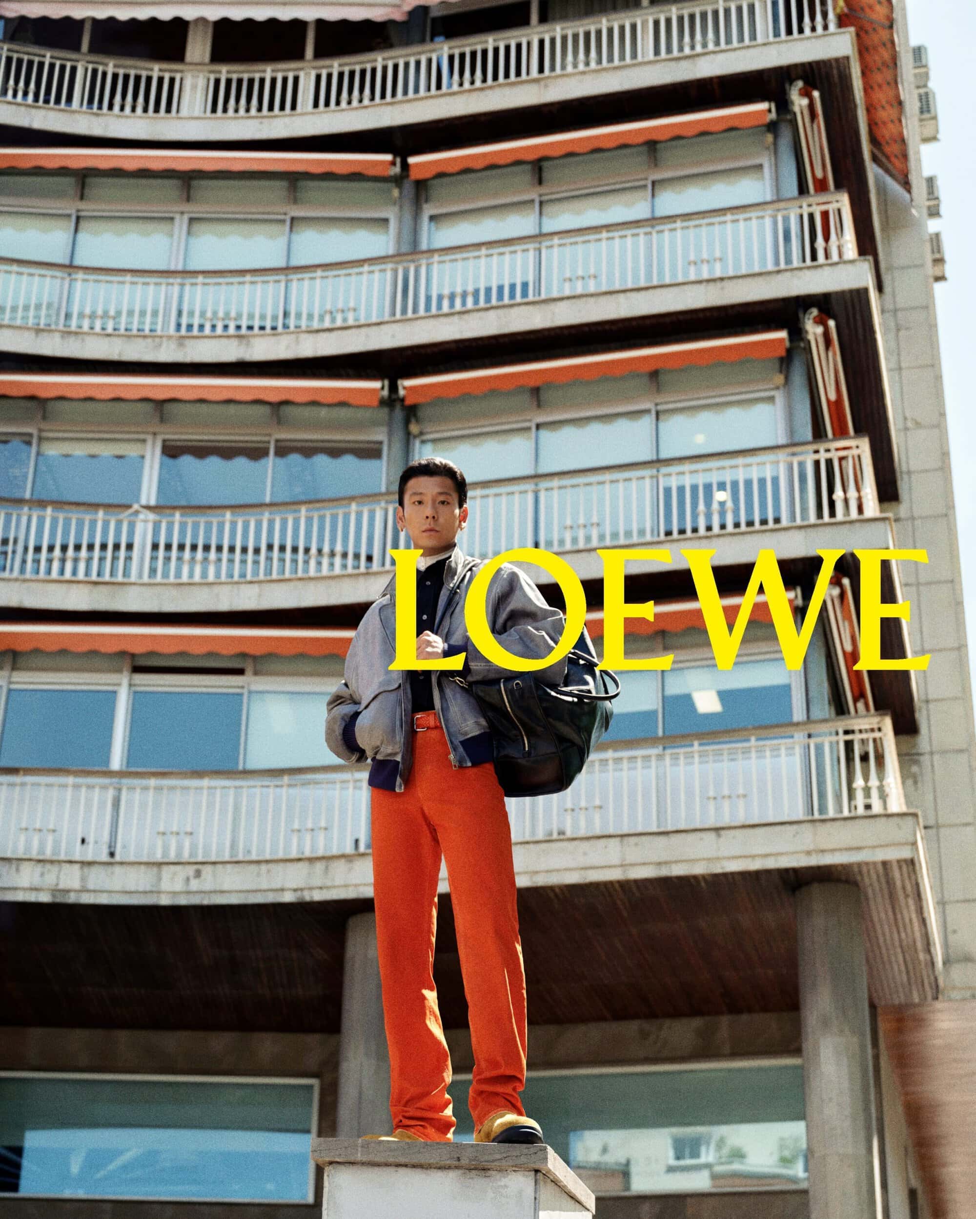 Man wearing a grey bomber jacket and bright orange trousers standing before a multi-story residential building facade with Loewe text overlay