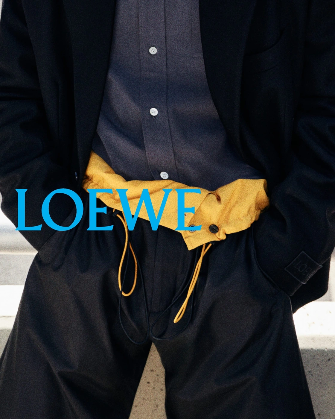 Close-up of person wearing a dark blazer, grey button-down shirt, mustard yellow waistband accent, and dark trousers with LOEWE overlay text.
