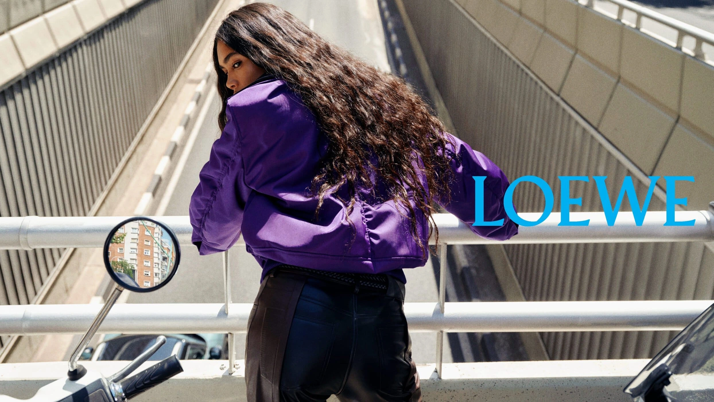 Model with long dark wavy hair wearing a purple jacket looking back from a motorcycle on an overpass with concrete barriers