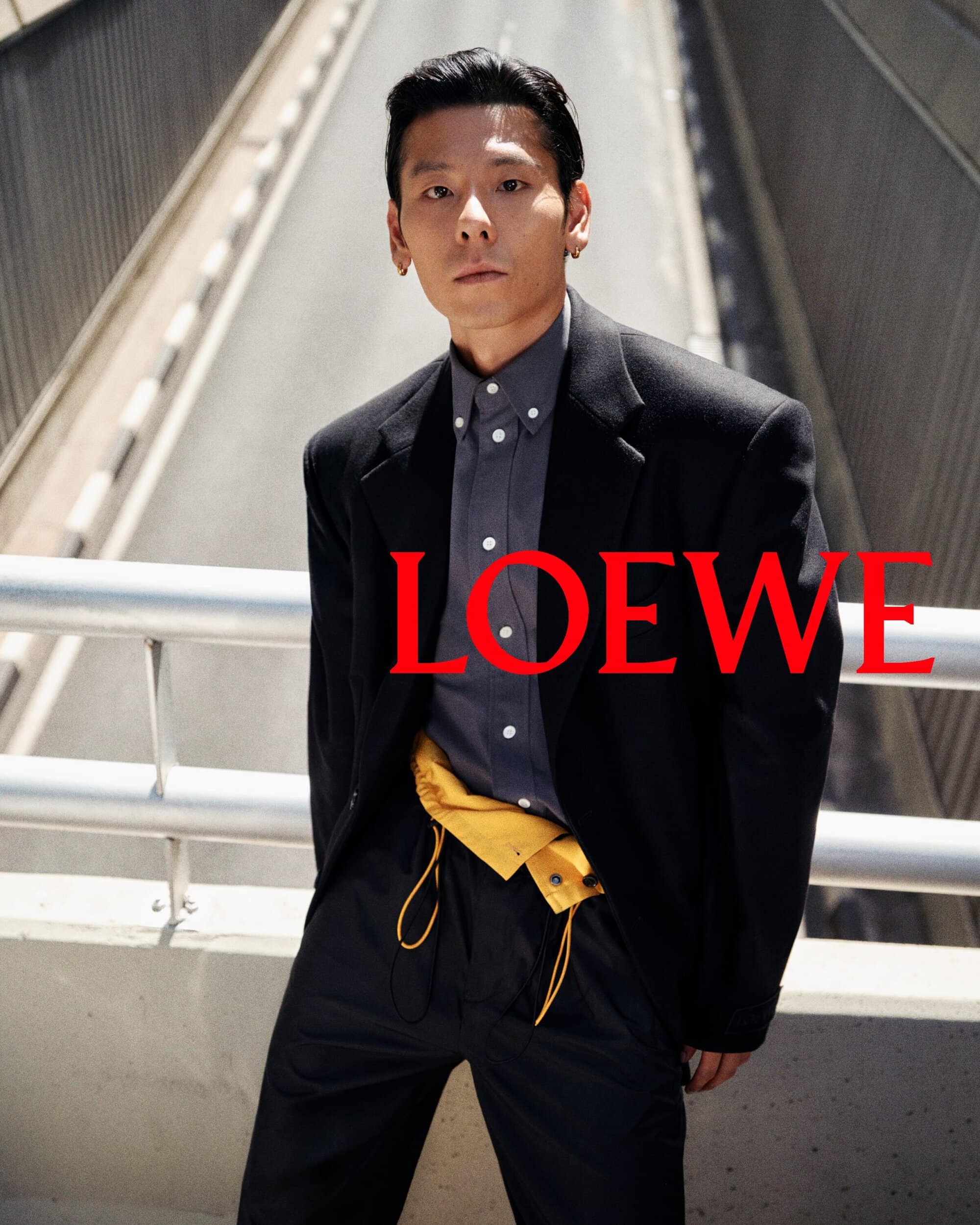 Man wearing a black blazer and grey button-up shirt standing on a concrete bridge for Loewe spring campaign