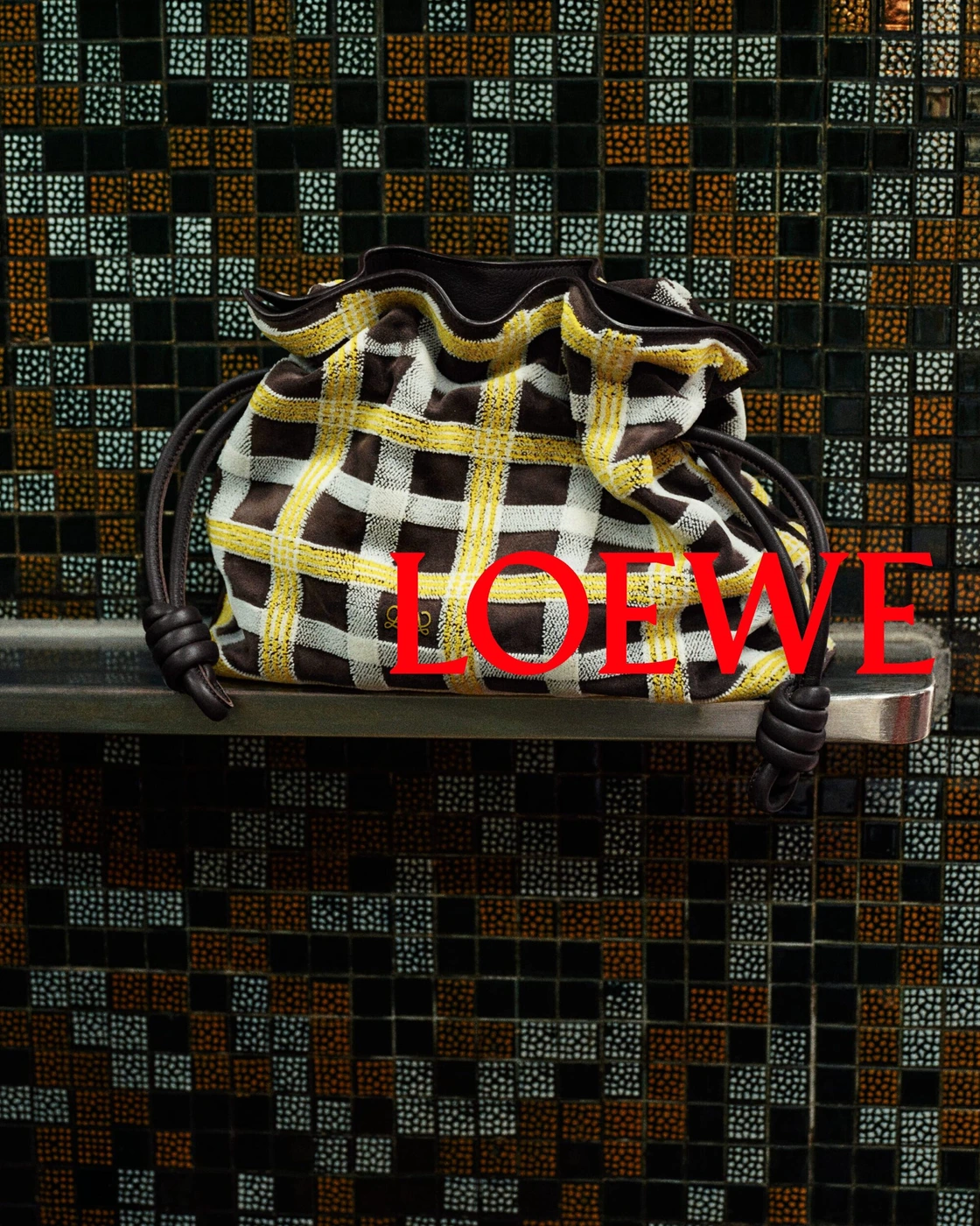 Loewe plaid drawstring handbag resting on a metal ledge against a dark mosaic tile wall