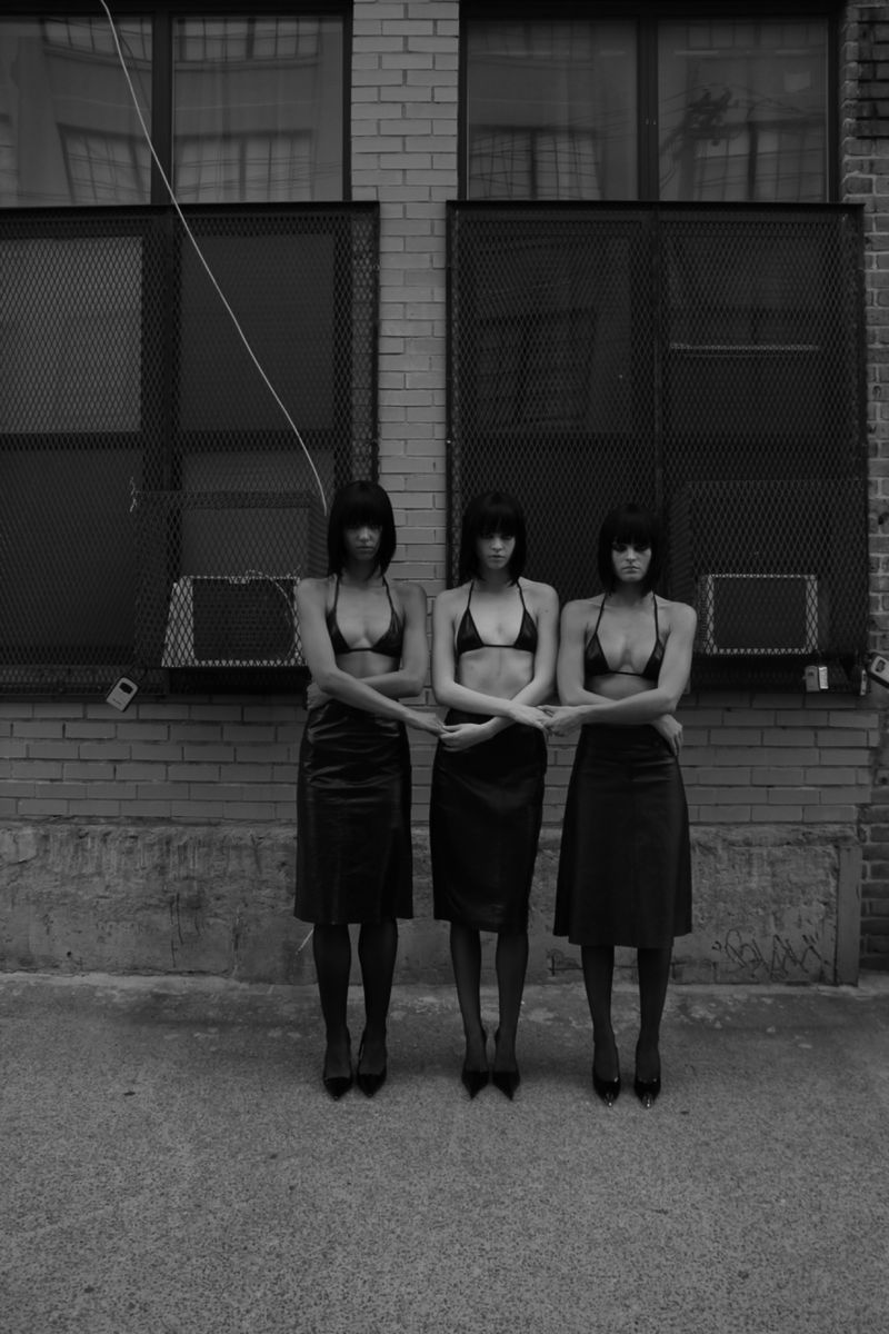 Three women stand outdoors in matching black tops and skirts, striking a pose with arms linked, against a textured brick wall backdrop.