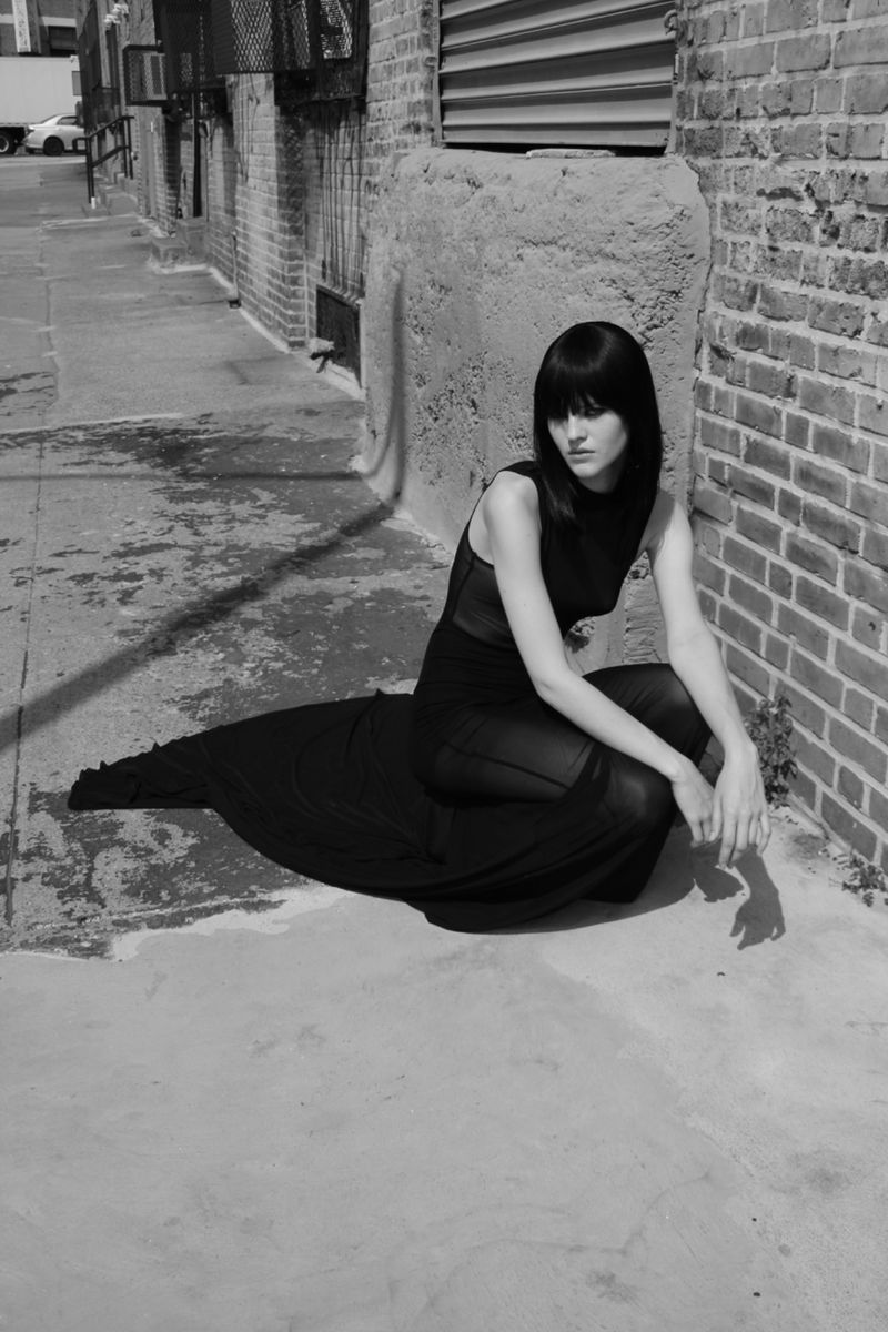 Monochrome outdoor fashion shot: a young woman in a black dress sits on a sidewalk next to a brick wall, bathed in bright sunlight.