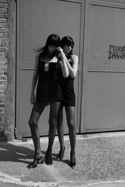 Black and white shot of two women in short black dresses and stockings, posing in front of a weathered metal gate in daylight.