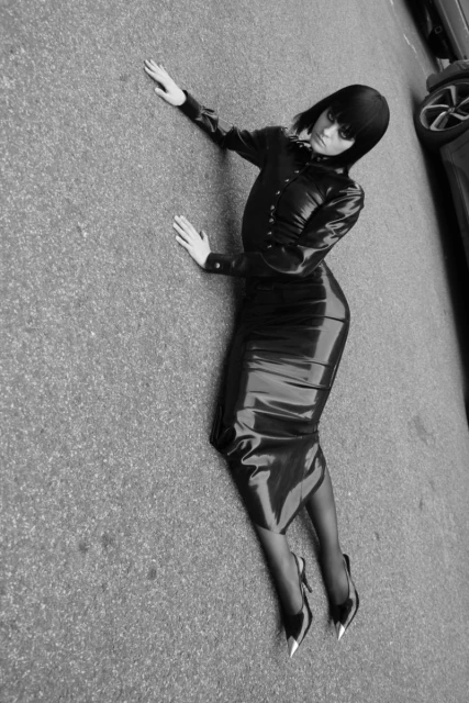 Black and white portrait of a woman in a latex outfit lying on pavement, featuring sleek style and a high-fashion aesthetic, with car in the background.