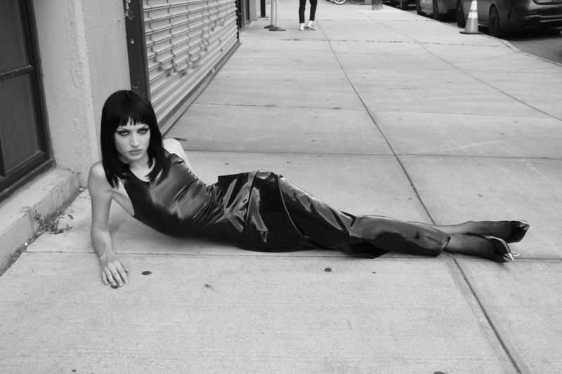 Monochrome studio shot of an adult woman in a dress lying on pavement, gazing at the camera with an enigmatic expression, urban setting.