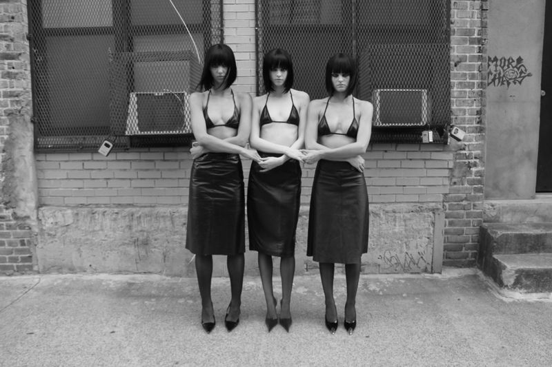 Three women stand arm in arm outside wearing matching black bikini tops, leather skirts, and heels in front of a brick wall.