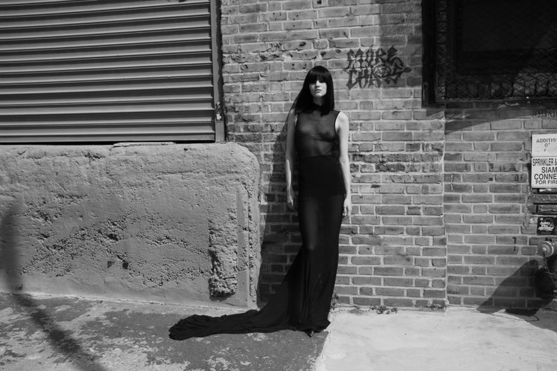 A slender adult female stands in a flowing black dress in front of a weathered brick wall and concrete structure in an outdoor urban setting.