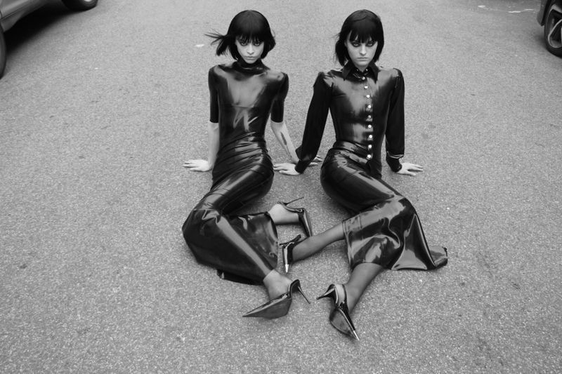 Two women in black glossy outfits and high heels sit on the asphalt in a monochrome fashion shot, showcasing a dark editorial style.
