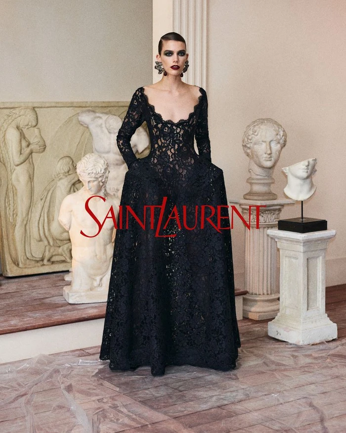 Model in a long black lace dress standing in a classic interior with classical sculptures and the Saint Laurent logo overlay.