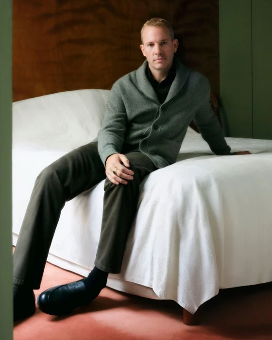 A man wearing a green cardigan and dark trousers sitting on a white bed in a wood-paneled room