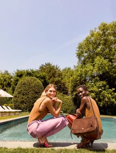 Two models squatting by an outdoor swimming pool wearing brown knit tops and trousers, featuring a brown suede shoulder bag.