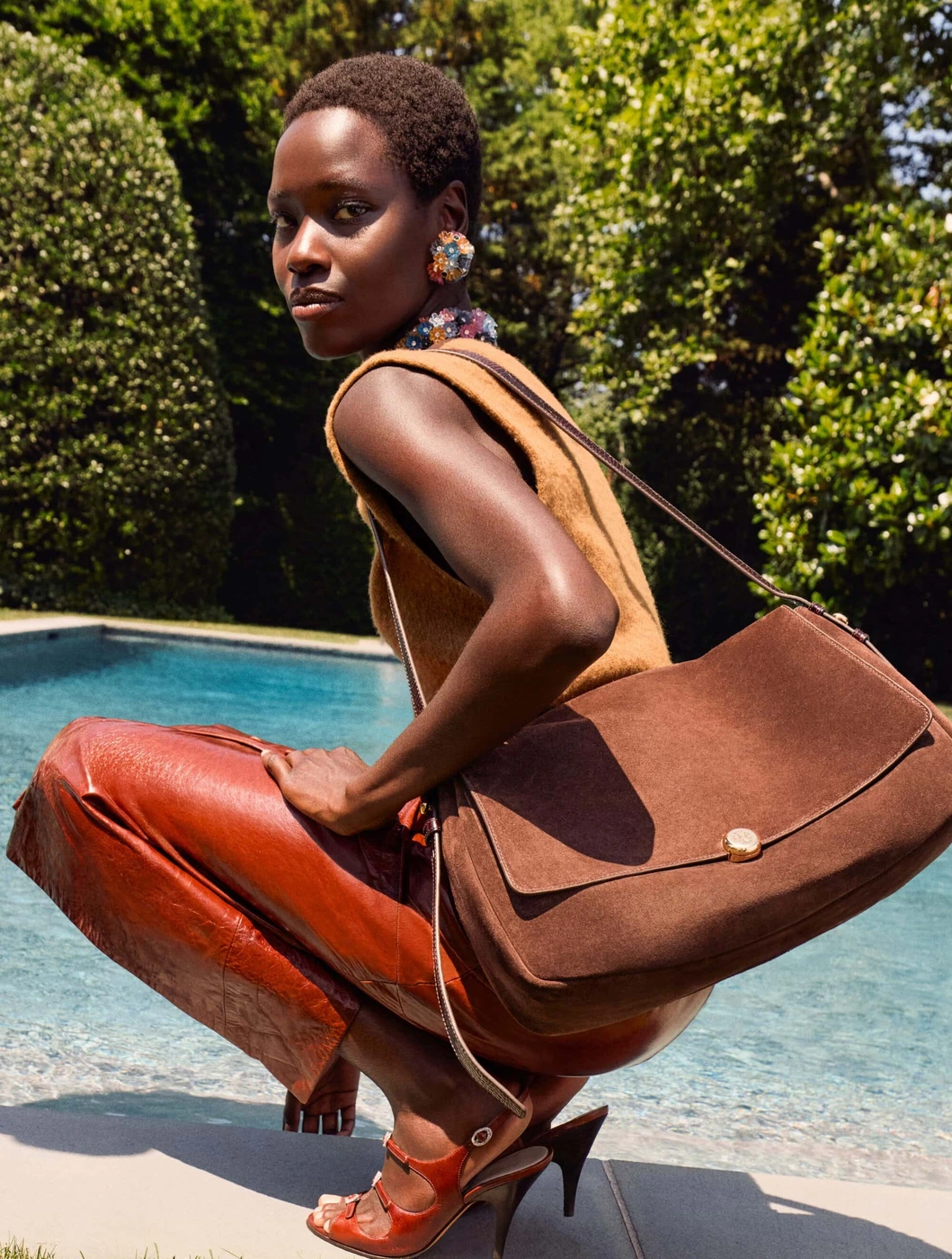 Model Awar Odhiang squatting by a swimming pool wearing a brown leather skirt, sleeveless top, and carrying a large brown suede shoulder bag.