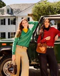 Two models leaning against a dark green off-road vehicle in front of a white house, wearing knit sweaters and carrying handbags.